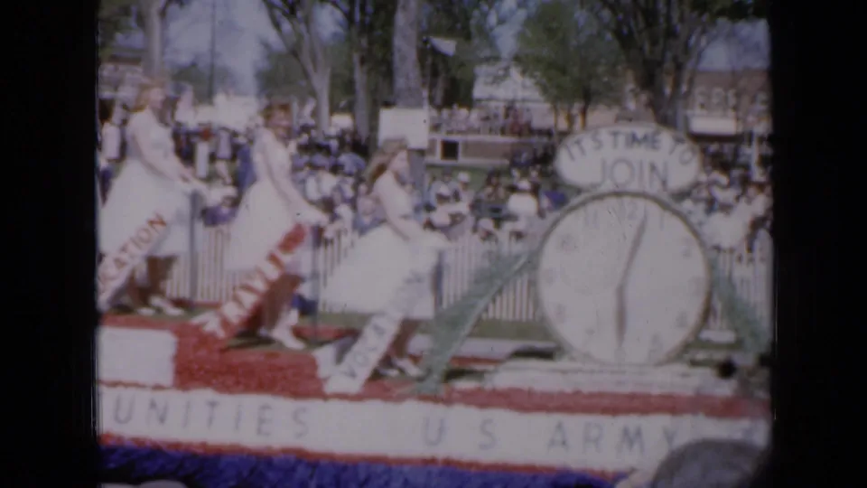 In 1959 Iowa home-movie footage, women in white dresses appear on a red, white, and blue U.S. Army recruitment float before spectators.