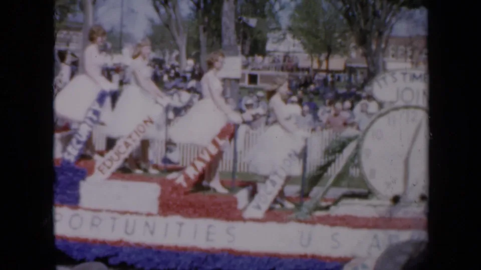 The Iowa parade float carries women in white dresses beside U.S. Army recruitment signs, bunting, spectators, and a large clock display.