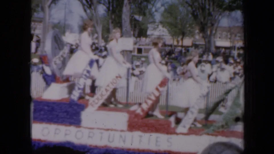 A home-movie frame shows the patriotic U.S. Army float with white-dressed riders, opportunity signage, and spectators along the parade route.