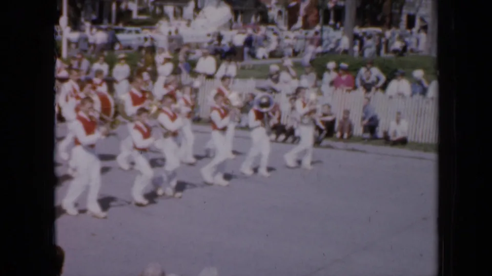 In the same 1959 Iowa parade, a red-and-white marching band plays while moving along a tree-lined street before seated and standing spectators.