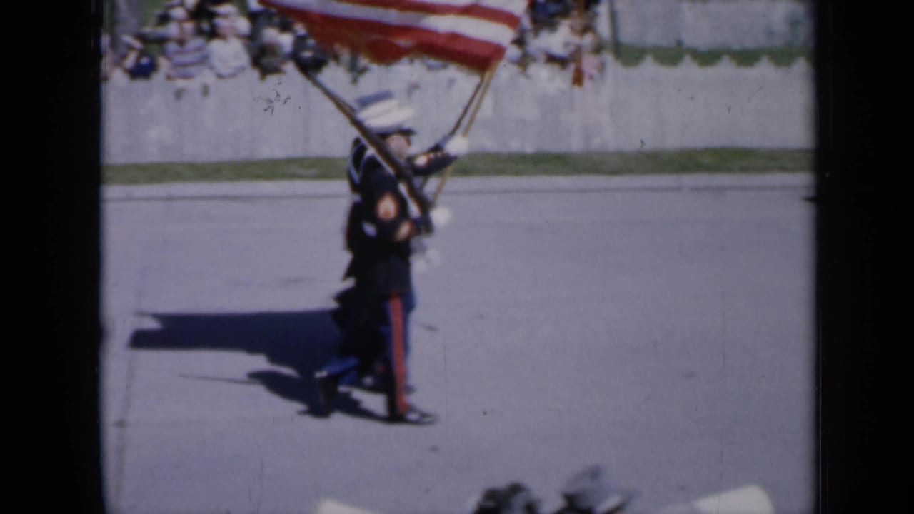 In 1959 Iowa home-movie footage, uniformed service members march with rifles and a large American flag during a parade.