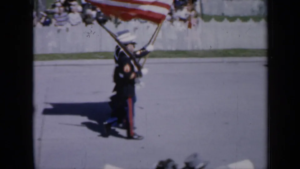 In 1959 Iowa home-movie footage, uniformed service members march with rifles and a large American flag during a parade.