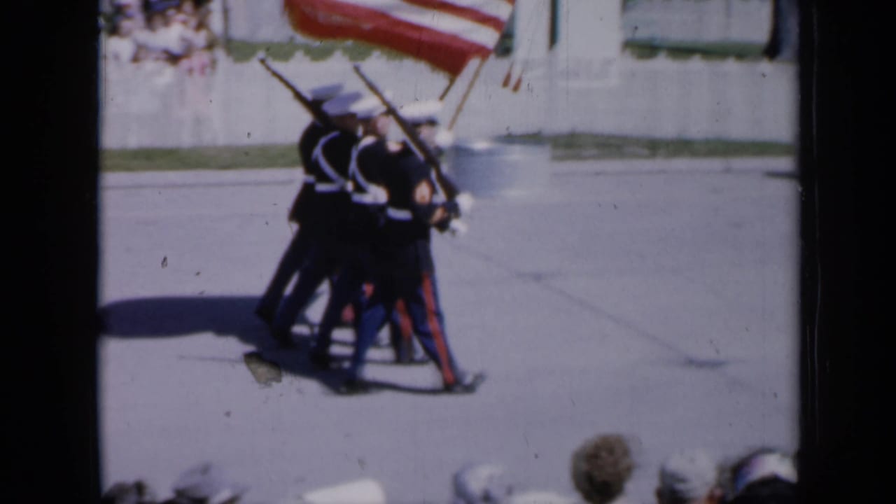 Honor guard members cluster while carrying rifles and a large American flag on the street.