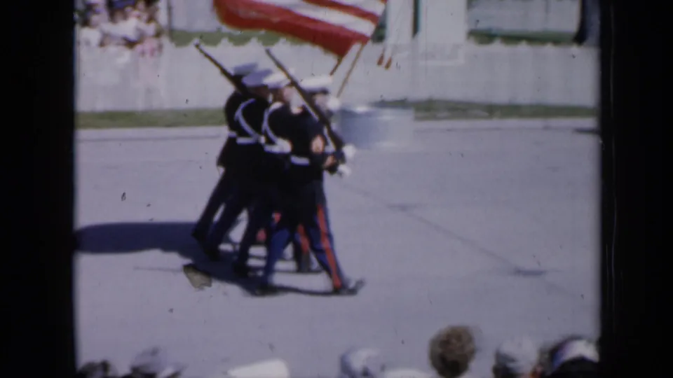 Uniformed honor guard members in Iowa appear mid-march with rifles and a large American flag, seen through soft 8mm color.