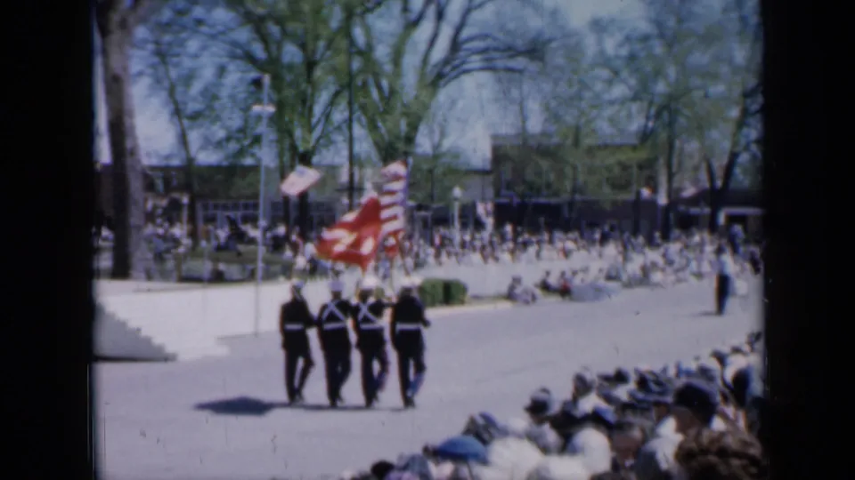 Four uniformed service members march away with a U.S. flag before a seated crowd in 1959 Iowa parade footage.