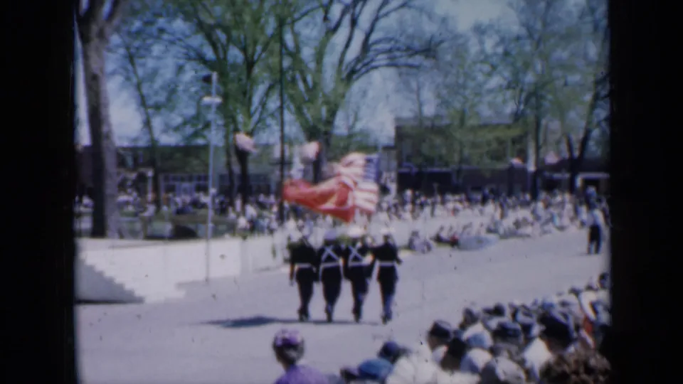 The honor guard marches away in 1959 Iowa footage, carrying a waving American flag past seated parade spectators.