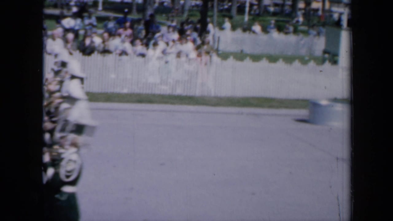 Marching band members enter the left edge of a sunlit street near spectators behind a white fence.