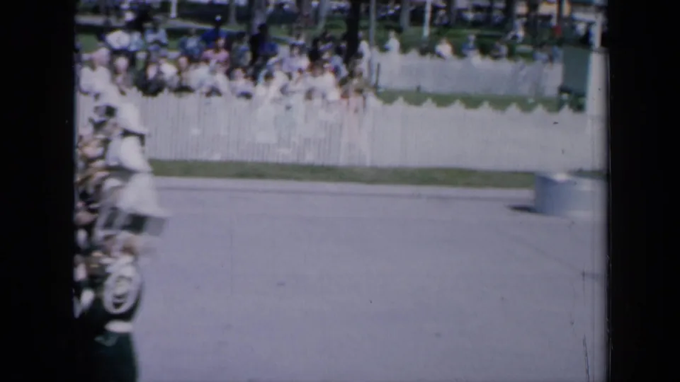 1959 Iowa home-movie frame showing band members partly visible at left as a parade moves along a bright street past a white fence and seated spectators.