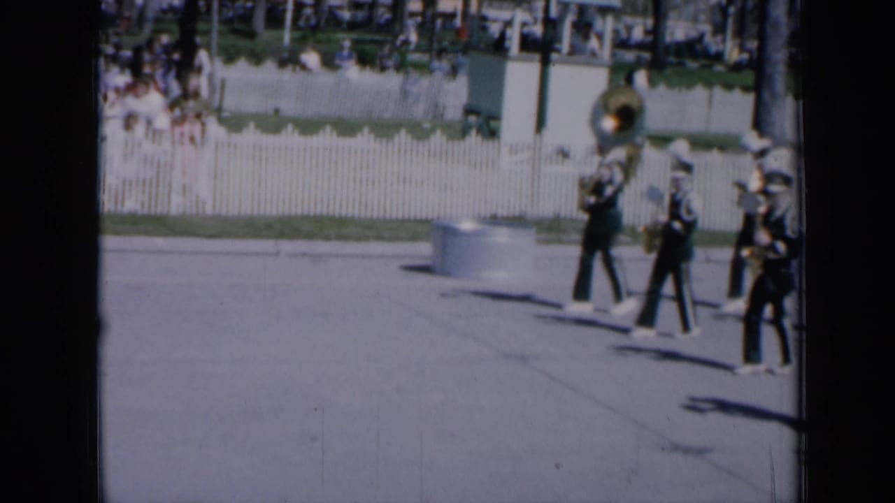 1959 Iowa parade frame with uniformed marching band members in white hats crossing a sunlit street, including a large brass instrument at rear.