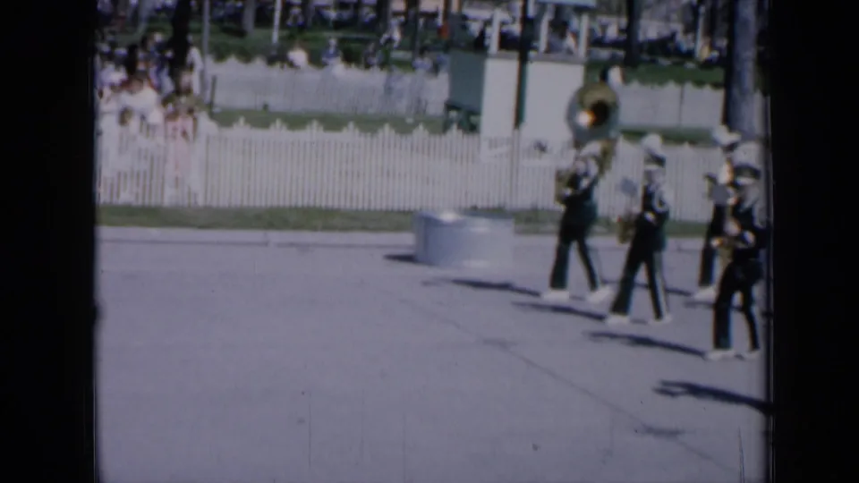 1959 Iowa parade frame with uniformed marching band members in white hats crossing a sunlit street, including a large brass instrument at rear.