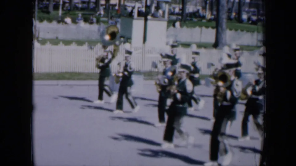 1959 Iowa home-movie frame showing a marching band in formation, with brass players and a large sousaphone moving along a street past a white fence.