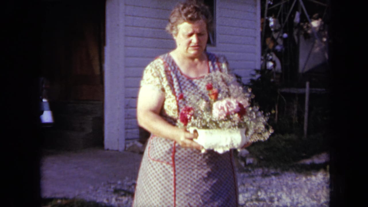 Woman in patterned apron holds a white vase of flowers outside a house.