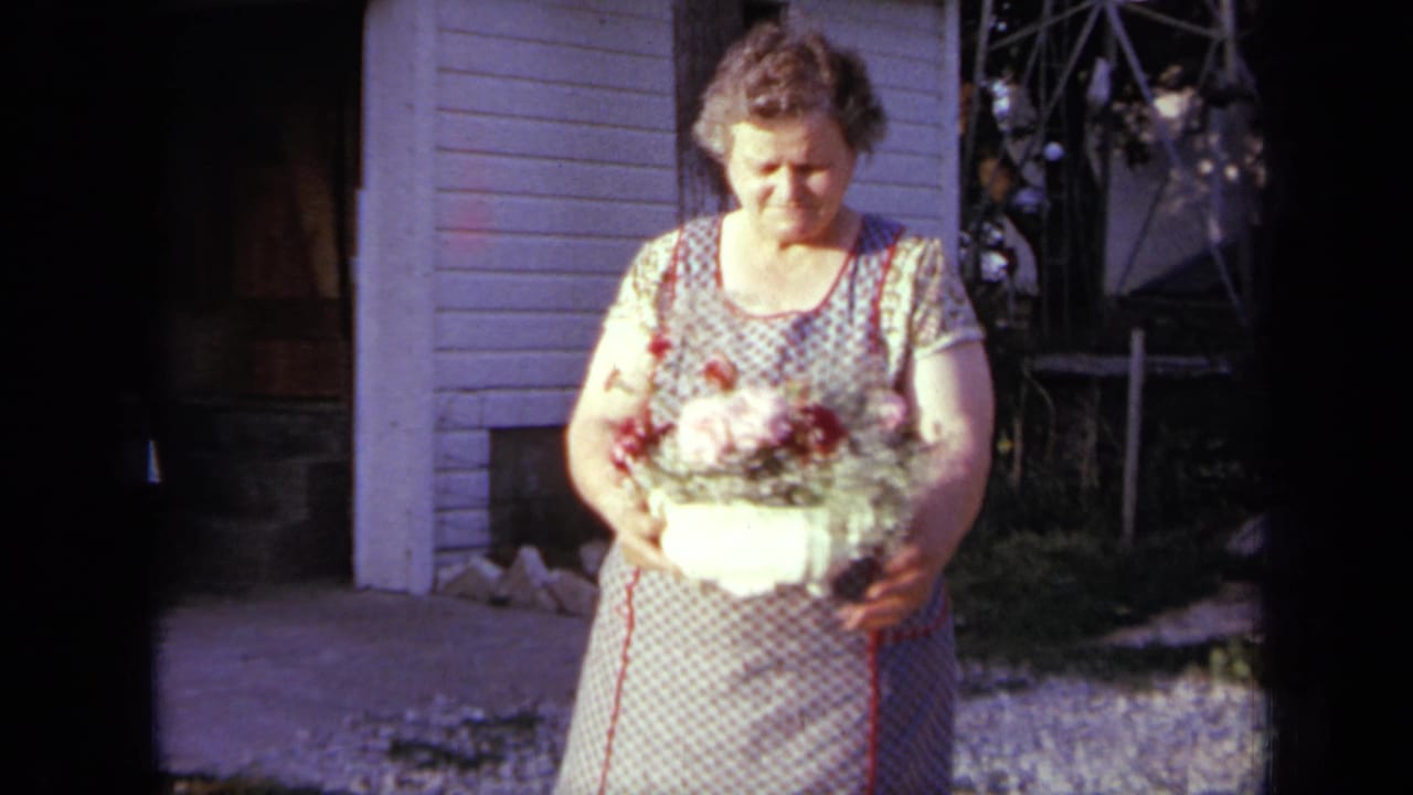 In Iowa, 1959, the woman looks down at the bouquet as she holds the white vase near the house exterior.