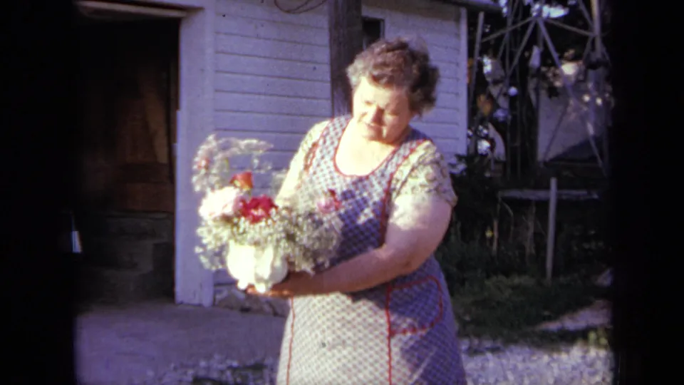 In Iowa, 1959, the woman adjusts or angles the flower arrangement in her hands beside the house doorway and window.