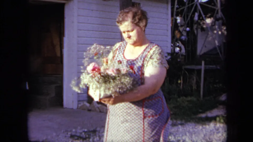 In Iowa, 1959, the woman stands in profile with the bouquet held outward, framed by house siding, a doorway, and a window.