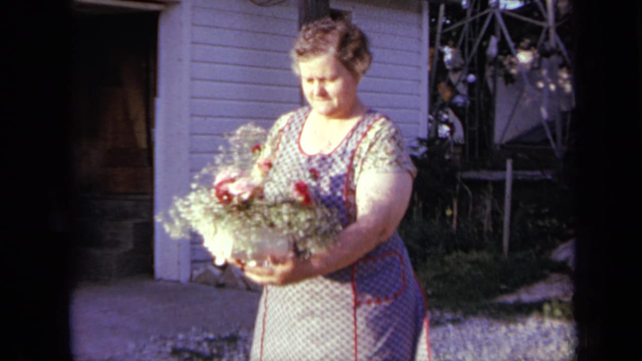 Older woman holding flowers outside a white-sided shed.