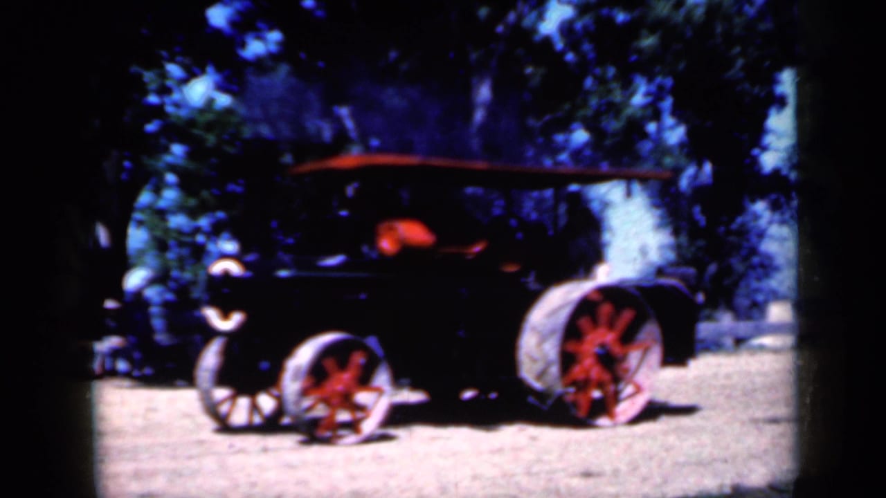 Black steam tractor with red-spoked wheels moving on dirt under trees.