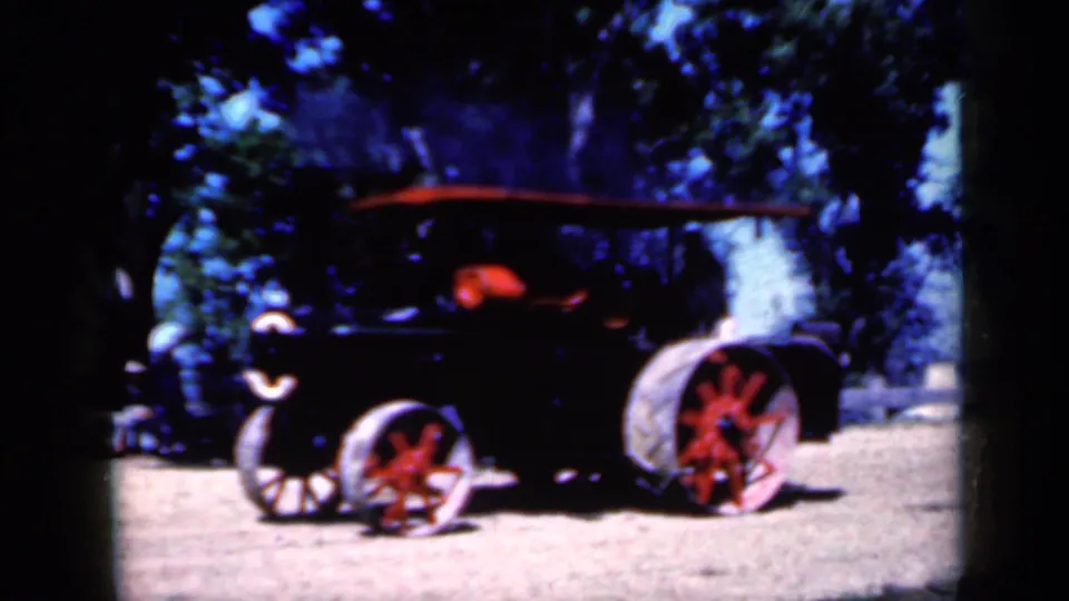 1959 Iowa 8mm frame of a black steam-powered tractor with red-spoked wheels crossing dirt ground beneath leafy trees.