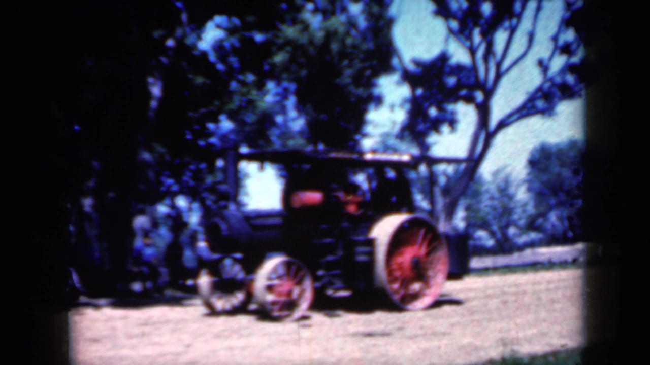 Side view of the Iowa steam tractor with red wheels and a visible operator as it passes through a tree-lined dirt area.