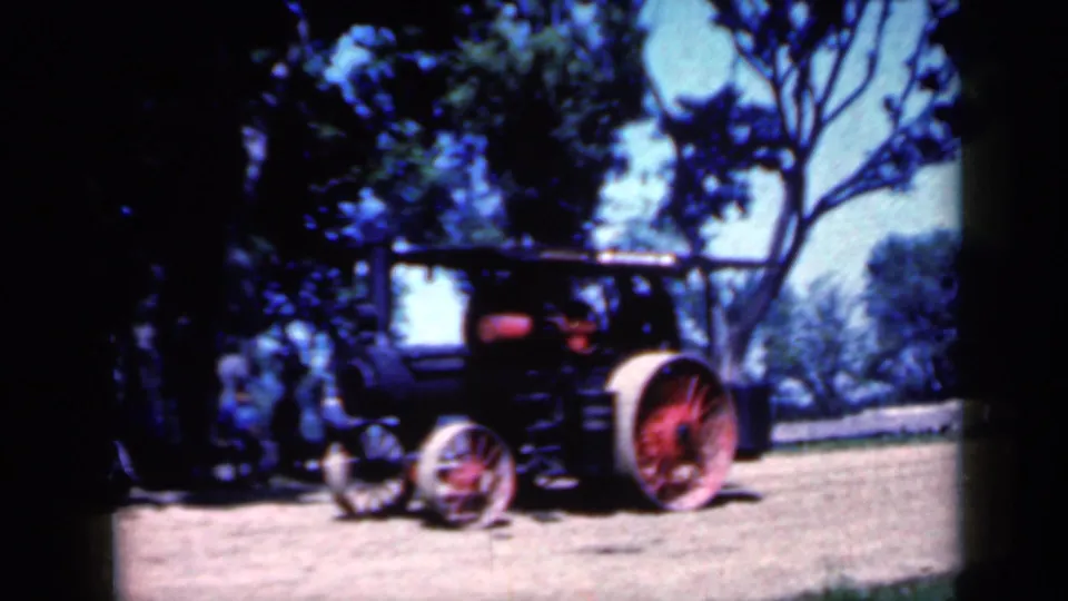 Side view of the Iowa steam tractor with red wheels and a visible operator as it passes through a tree-lined dirt area.
