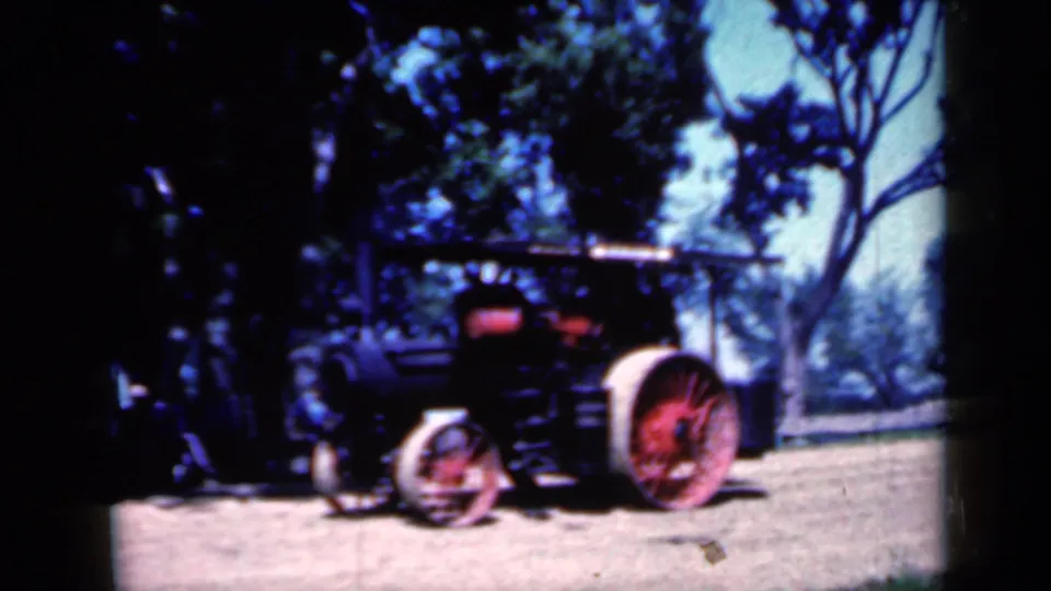A 1959 Iowa home-movie frame shows the steam tractor in partial shade, its red-spoked wheels standing out against the dirt ground.