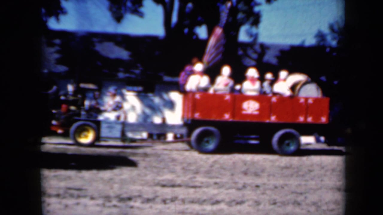 Red passenger wagon with seated visitors beside a black steam tractor on a dirt fairground.