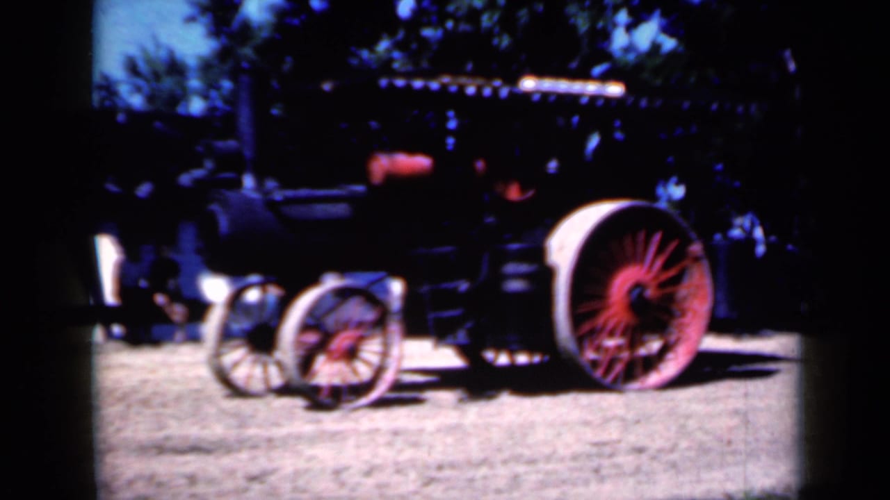1959 Iowa home-movie frame of a black steam tractor with a large red-spoked rear wheel and pale rim on a shaded dirt fairground.