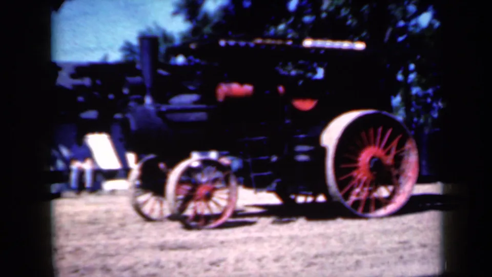 1959 Iowa frame showing the side of a black steam tractor, its red-spoked rear wheel, smaller front wheels, and strong sunlight on the dirt ground.
