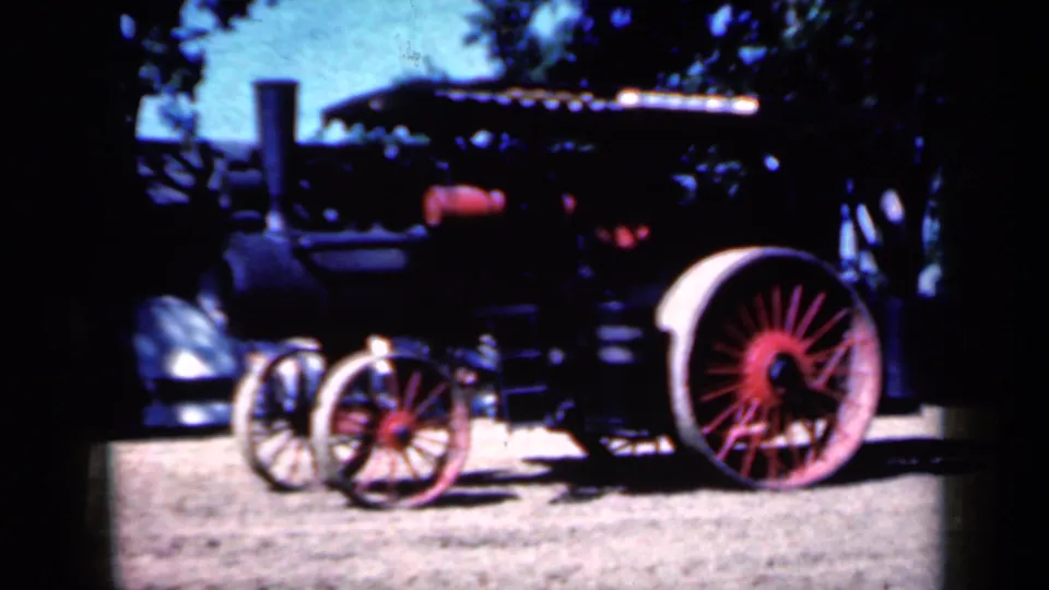 1959 Iowa home-movie frame of the steam tractor from the side, with red-spoked wheels, black machinery, dirt ground, and trees in the background.