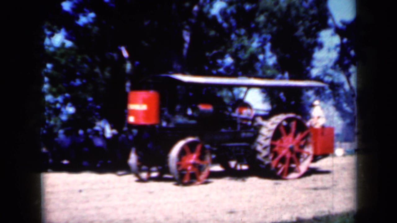 1959 Iowa home-movie frame showing a black steam traction engine with red spoked wheels, a red tank, and a canopy on a dirt field.