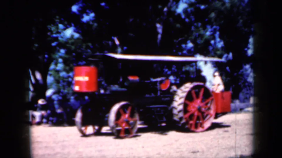 Closer 1959 Iowa frame of the steam traction engine, emphasizing the red spoked wheels, black body, red tank, and shaded tree backdrop.