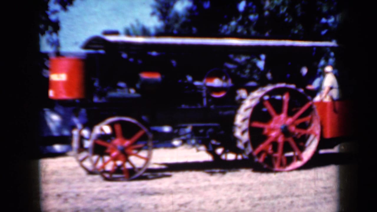 Black steam tractor with red spoked wheels parked on dirt under trees.