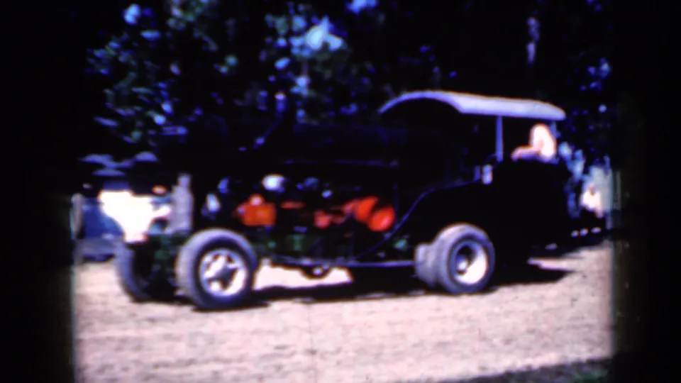 1959 Iowa 8mm frame showing a dark open-top vehicle or machinery beside the steam equipment area, with red parts and a seated person visible.