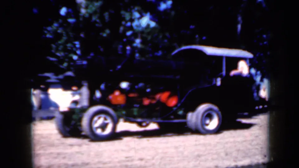 1959 Iowa 8mm frame showing a black canopy-covered steam tractor on dirt ground with trees behind it.