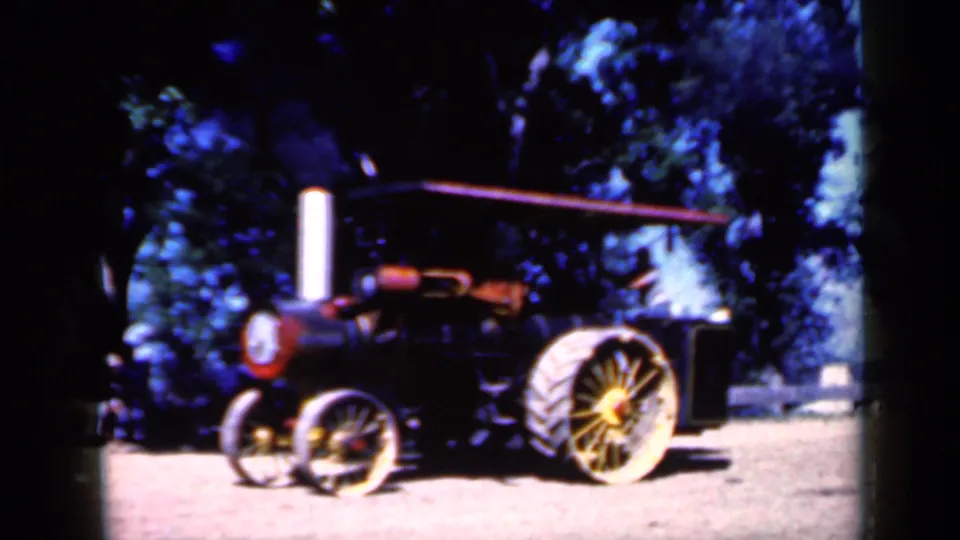 1959 Iowa home-movie frame showing a red and black steam traction engine with yellow-spoked wheels parked beneath shade trees.