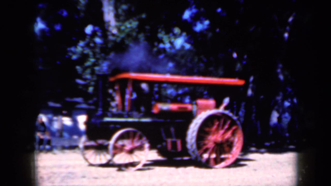 Steam-powered tractor with red spoked wheels on a dirt road under trees.