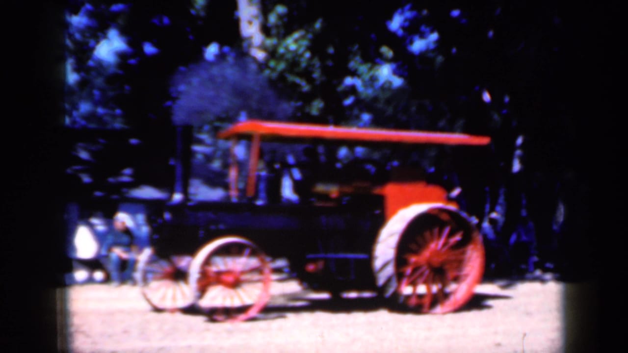 Steam tractor seen side-on with red wheels, canopy, trees, and people nearby.