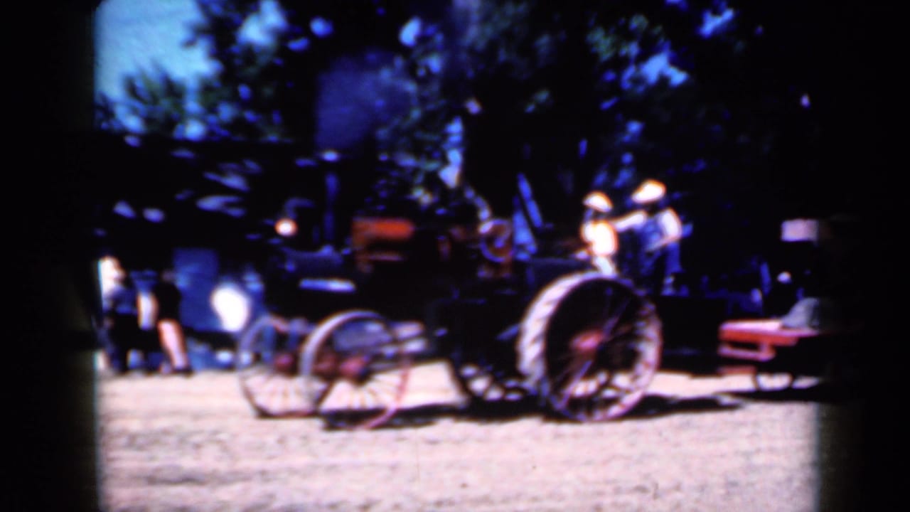 1959 Iowa home-movie frame showing a black steam tractor with red wheels at a rural fair setting, with people and cars nearby.