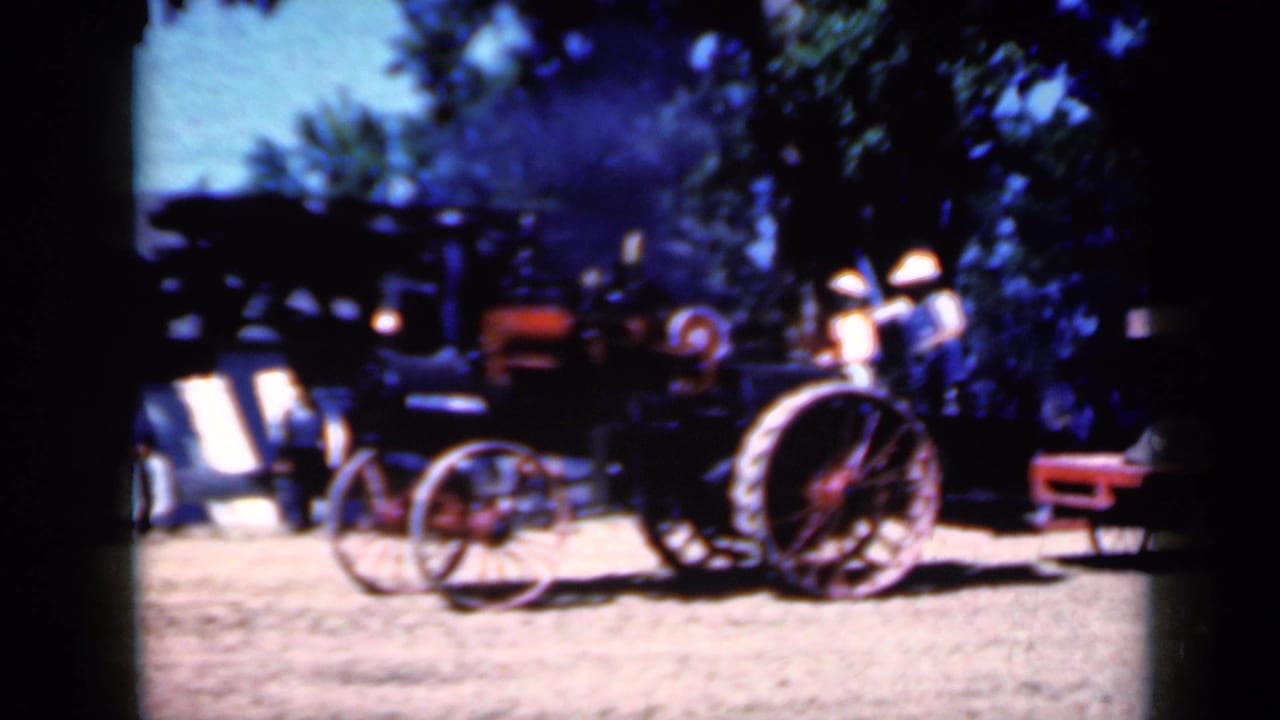Side view of a vintage steam tractor at an Iowa rural fair, with red wheels, shaded trees, and people standing around it.
