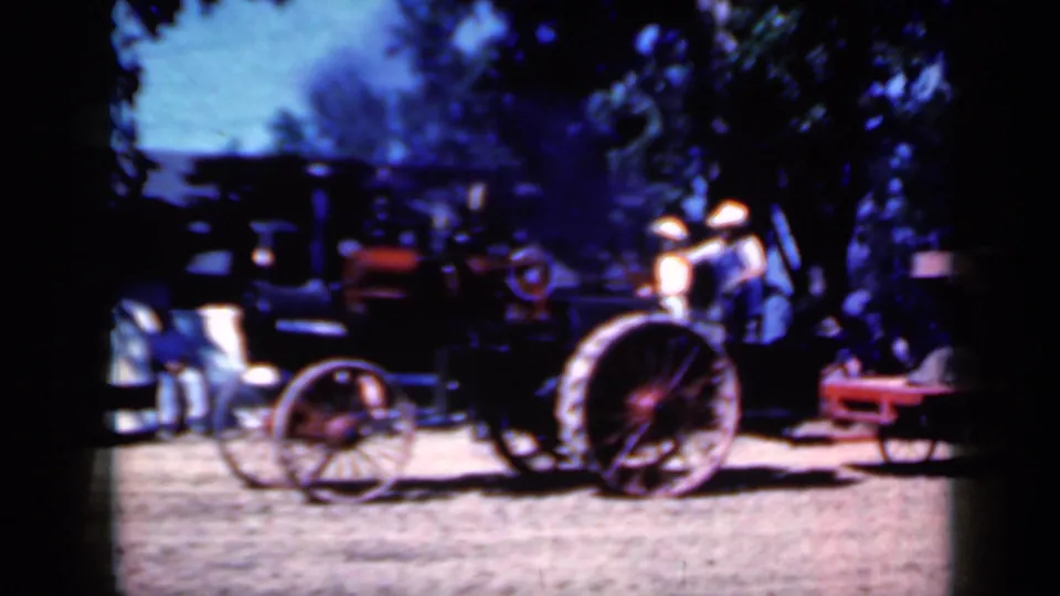 Closer 8mm view of the steam tractor's large red wheel and black body, with fair visitors and cars in the background.