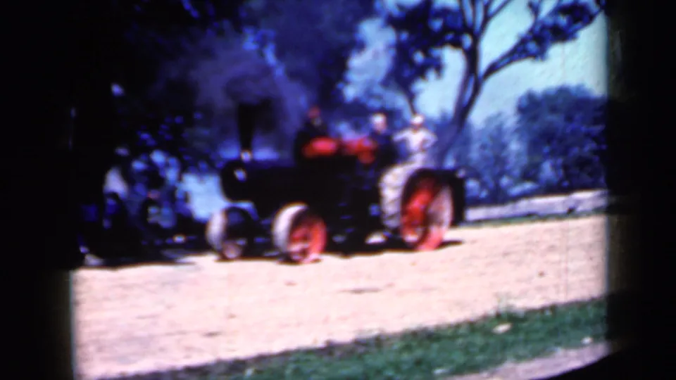 Steam tractor with red wheels and canopy seen farther along a dirt road in rural Iowa, bordered by grass and trees.