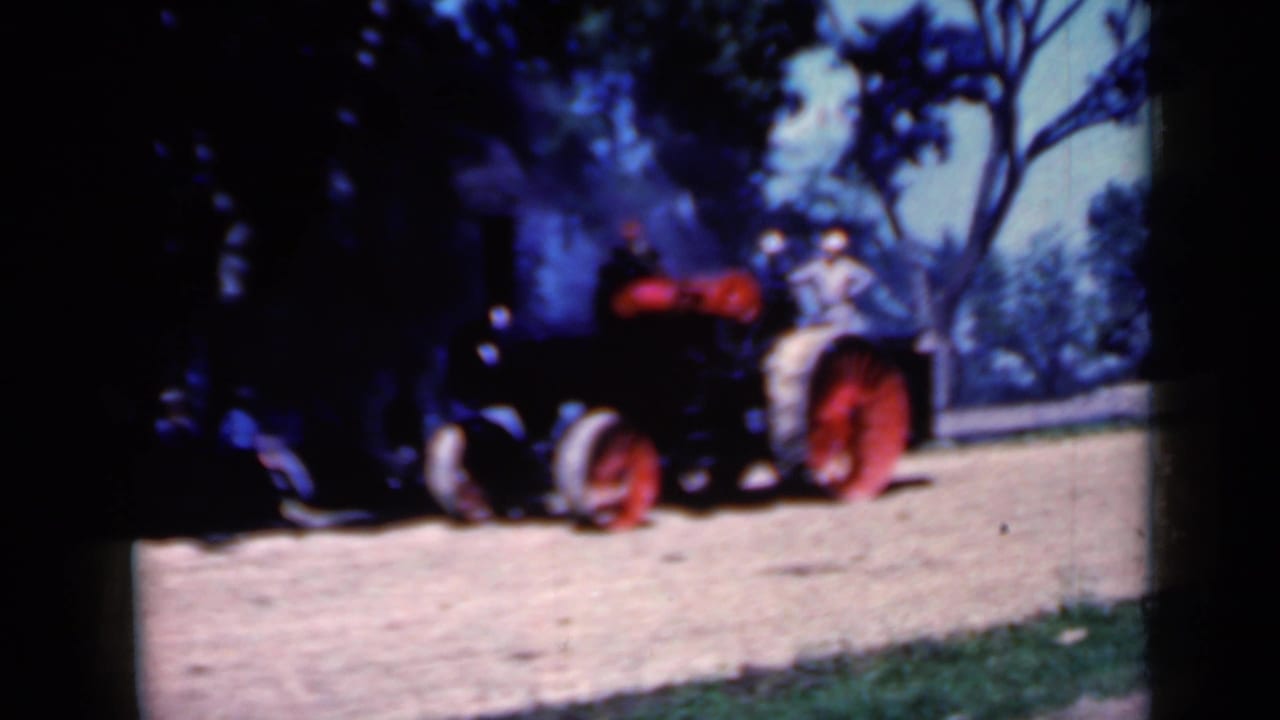The Iowa farm tractor continues toward the camera, carrying two visible riders as smoke drifts upward in the tree-lined background.