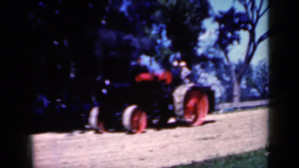 Near the end of the clip, the red-wheeled tractor is close to the camera, with riders aboard and smoke rising against the trees.