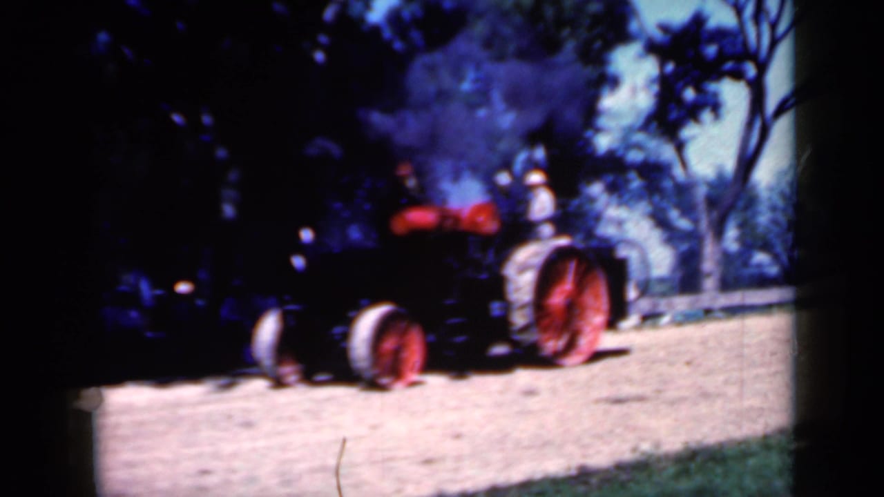 1959 Iowa 8mm frame showing a black steam tractor with red wheels crossing sunlit dirt as smoke drifts upward.
