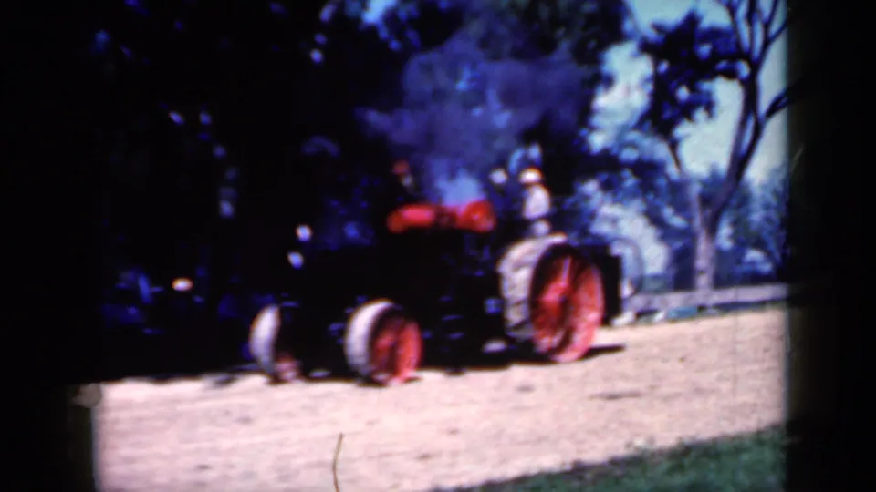 1959 Iowa 8mm frame showing a black steam tractor with red wheels crossing sunlit dirt as smoke drifts upward.
