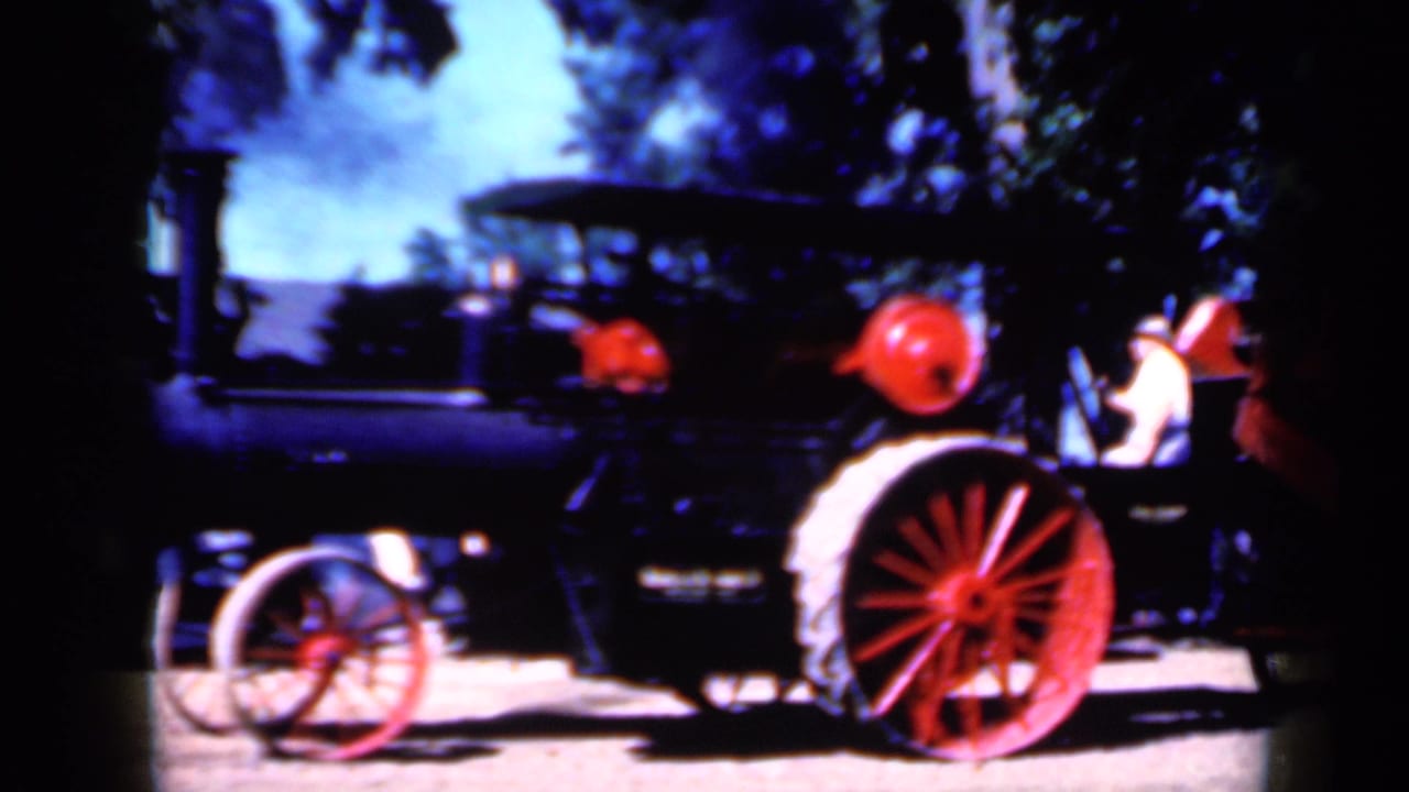 Red agricultural machine beside a dark older car with adults nearby outdoors.