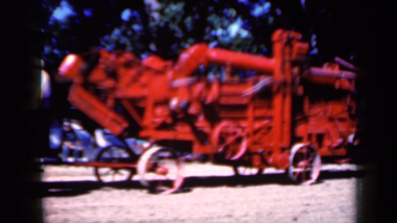 Large red agricultural machine with wheels and belts parked under trees.