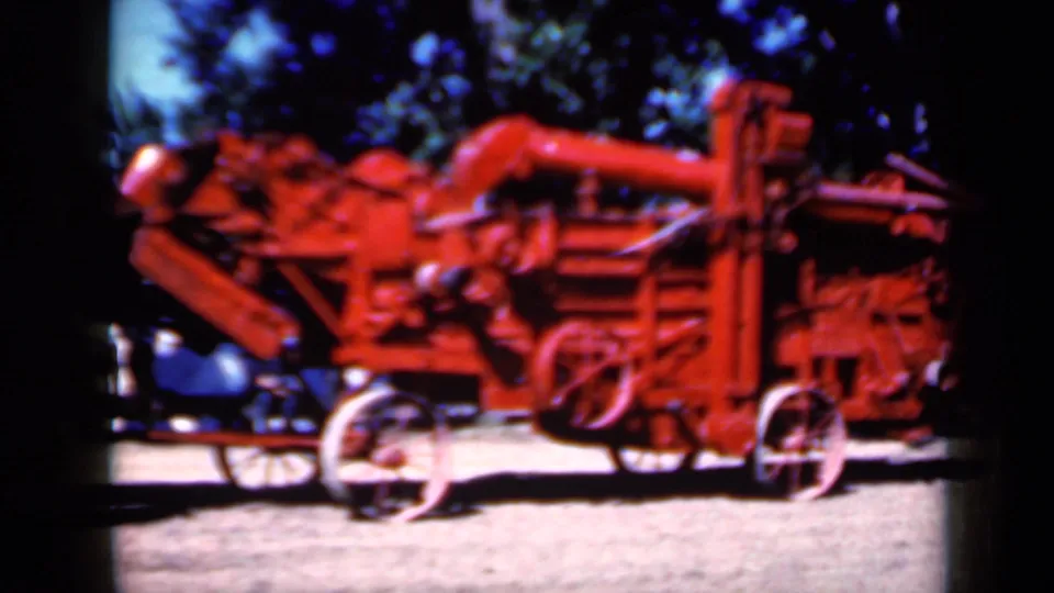 Close 8mm frame of the red agricultural machine, showing spoked wheels, belt assemblies, and a dark older car partly behind it.