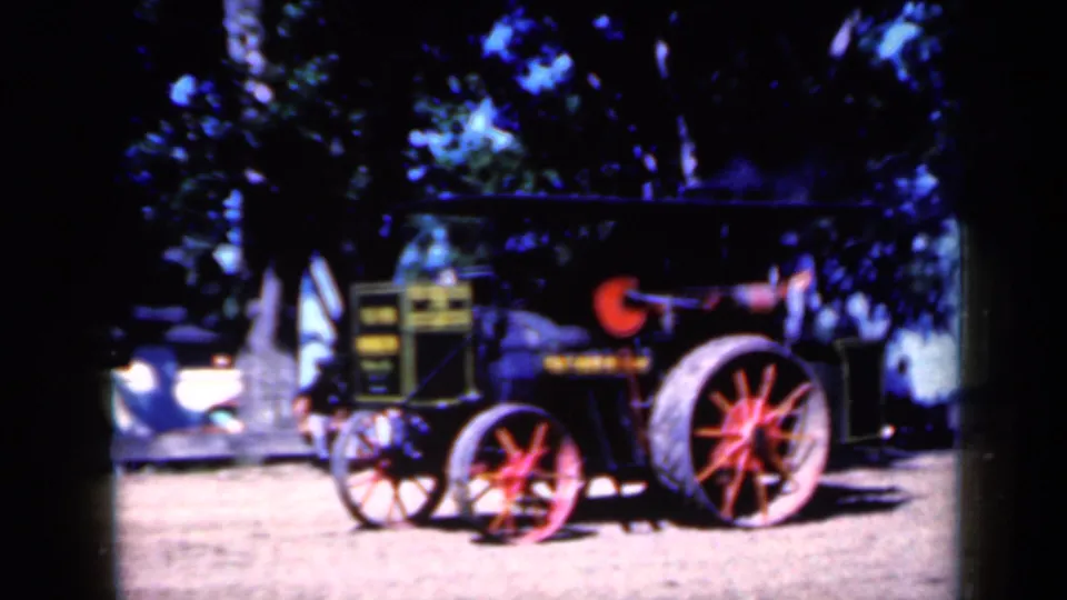 Closer 8mm view of the vintage tractor's red-spoked wheels and dark machinery in an Iowa rural event setting.