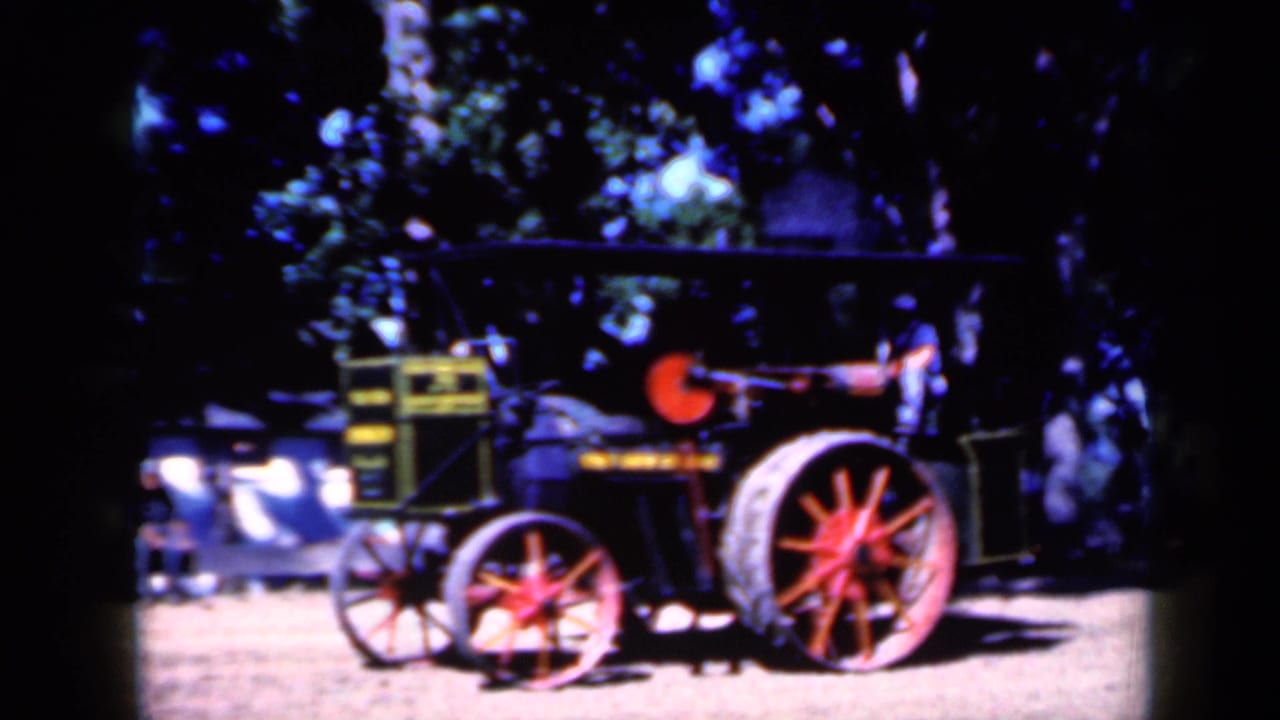 Black steam engine with red wheels moving on a dirt field under trees.